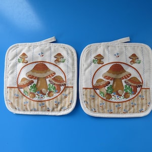 May include: Two square pot holders featuring a mushroom design on a white background. The mushrooms are brown and tan with green leaves. Each pot holder has a loop for hanging.