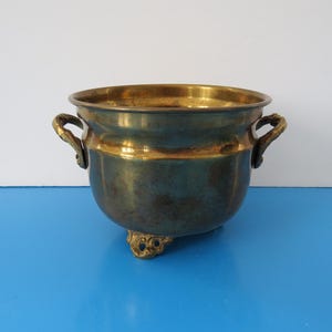 May include: A brass planter with two handles and three feet. The planter has a worn, aged look.