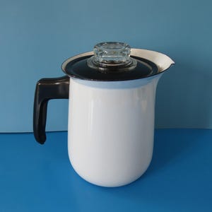 May include: A white enamel coffee pot with a black handle and lid. The lid has a clear glass knob. The pot has a curved shape and a spout for pouring. The background is a solid blue.