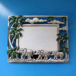 Peut inclure: Un cadre photo rectangulaire en métal représentant une scène de jungle avec trois éléphants argentés marchant dans l'herbe. Le cadre est orné de palmiers émaillés verts et de nuages blancs sur le dessus et les côtés, entourant une ouverture centrale.