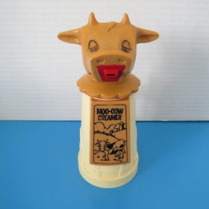 Peut inclure: Un ancien distributeur de crème Moo-Cow, un distributeur de crème fantaisie. Le distributeur a la forme d'une tête de vache avec les yeux fermés et une bouche rouge. La base est de couleur jaune clair avec une étiquette qui dit "MOO-COW CREAMER".
