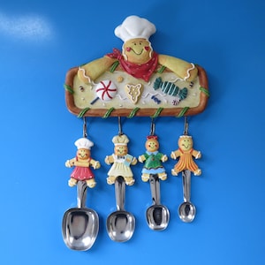 Peut inclure: Un support à ustensiles de cuisine mural avec une figurine de chef en pain d'épices en haut. Quatre cuillères à mesurer en métal sont suspendues à des crochets, chacune surmontée d'un petit personnage en pain d'épices vêtu de vêtements colorés.