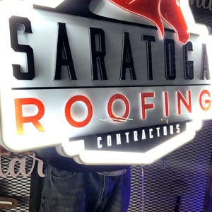 Puede incluir: Letrero luminoso para Saratoga Roofing Contractors. El letrero presenta el nombre de la empresa en letras negras en negrita, con "ROOFING" en rojo. Un gráfico de cabeza de caballo rojo está en la parte superior. El letrero es blanco con un borde blanco brillante.