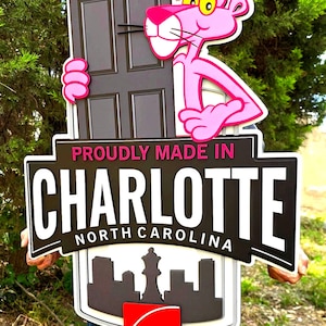 May include: A rectangular sign featuring the Pink Panther character holding a dark door. The sign displays the text PROUDLY MADE IN CHARLOTTE NORTH CAROLINA above a black city skyline silhouette and a red Owens Corning logo at the bottom.