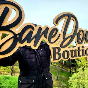 May include: A wooden sign with the words "Bare Down Boutique" in black lettering. The sign is held in front of a person wearing a black jacket.