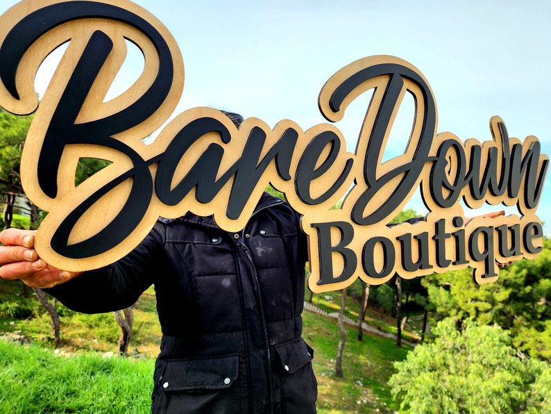 May include: A wooden sign with the words "Bare Down Boutique" in black lettering. The sign is held in front of a person wearing a black jacket.