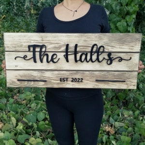 May include: A rustic wooden sign with the text "The Halls" in black cursive lettering and "EST 2022" below. The sign is held by a person in front of a green leafy background.