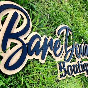 May include: A wooden sign with the text "Bare Down Boutique" in black lettering. The sign is on a bed of green grass.