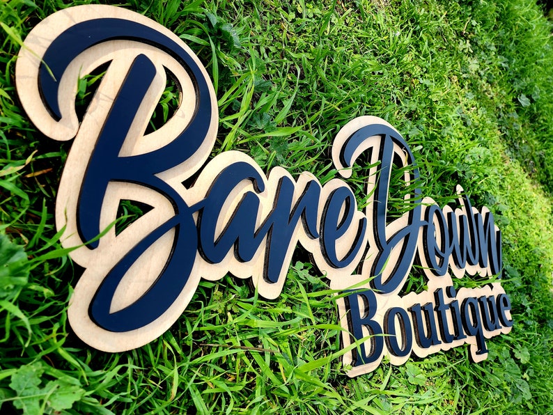 May include: A wooden sign with the text "Bare Down Boutique" in black lettering. The sign is on a bed of green grass.