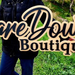 May include: A wooden sign with the text "Bare Down Boutique" in black lettering. The sign is held by a person wearing a black jacket and blue jeans.