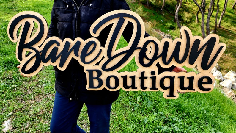 May include: A wooden sign with the text "Bare Down Boutique" in black lettering. The sign is held by a person wearing a black jacket and blue jeans.