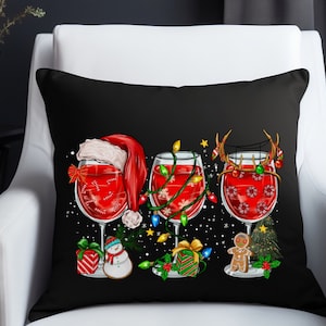 May include: A black throw pillow with a Christmas-themed design featuring three wine glasses filled with red wine. Each glass has a different festive topper: a Santa hat, a string of lights, and reindeer antlers. The glasses are surrounded by Christmas decorations, including a snowman, a gingerbread man, and a Christmas tree.