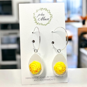 May include: Silver hoop earrings with a white egg-shaped base and a yellow yolk design with orange speckles. The earrings are displayed on a white card with the text "jo-lea Muse" and "HANDMADE JEWELRY KEEPSAKES AND ADORNMENTS."