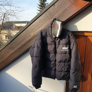 Può includere: Una giacca imbottita nera con una cerniera bianca e un logo Helly Hansen bianco sul davanti. La giacca è appesa a un appendiabiti contro un muro di legno.