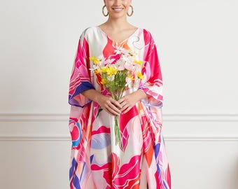 May include: A vibrant, flowing caftan dress with a V-neckline. The dress features a colorful abstract floral pattern in shades of pink, red, orange, and blue. The model is holding a bouquet of flowers. The dress is floor-length.