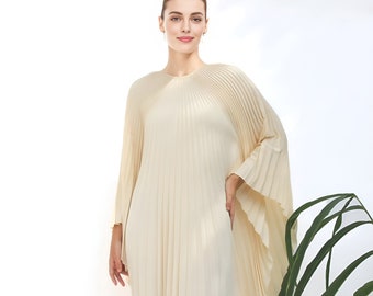 May include: A woman wearing a long, cream-colored pleated dress with a flowing, wide-sleeved design. The dress has a simple, round neckline and is made of a lightweight, flowing fabric.