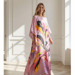 May include: A flowing, pink caftan dress with a colorful abstract floral design. The dress features a round neckline, wide sleeves, and a floor-length hemline. The pattern includes yellow, black, and purple accents. The model is wearing high heels.