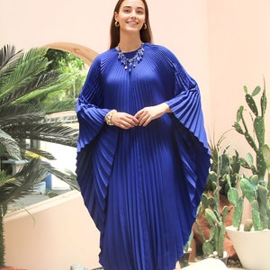 May include: A woman wearing a royal blue pleated maxi dress with a blue beaded necklace and white heels. She is standing in front of a pink wall with plants in the background.
