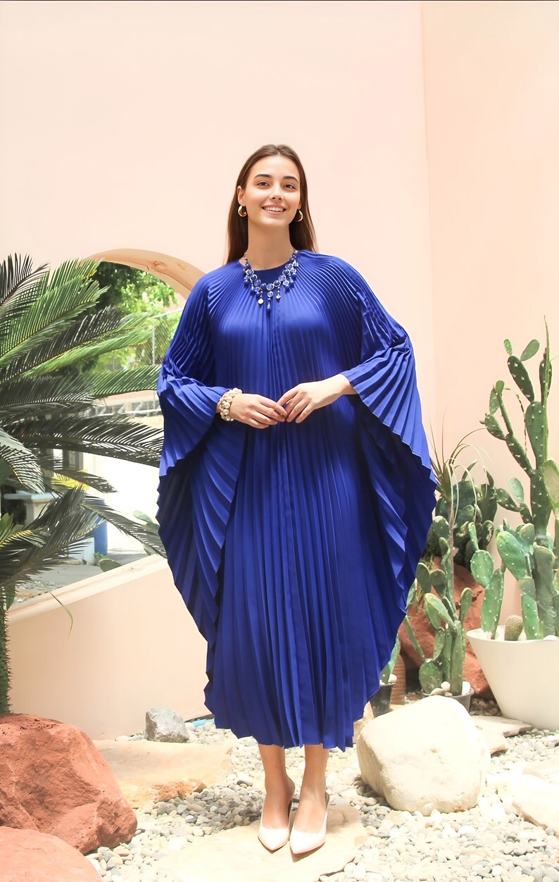 May include: A woman wearing a royal blue pleated maxi dress with a blue beaded necklace and white heels. She is standing in front of a pink wall with plants in the background.