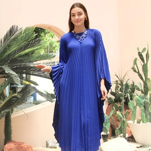 May include: A royal blue pleated maxi dress with long sleeves and a round neckline. The dress has a flowing, relaxed fit and features a decorative beaded necklace.