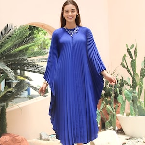May include: A woman wearing a royal blue pleated maxi dress with long sleeves and a silver necklace. She is standing in front of a pink wall with a cactus and a palm tree in the background.