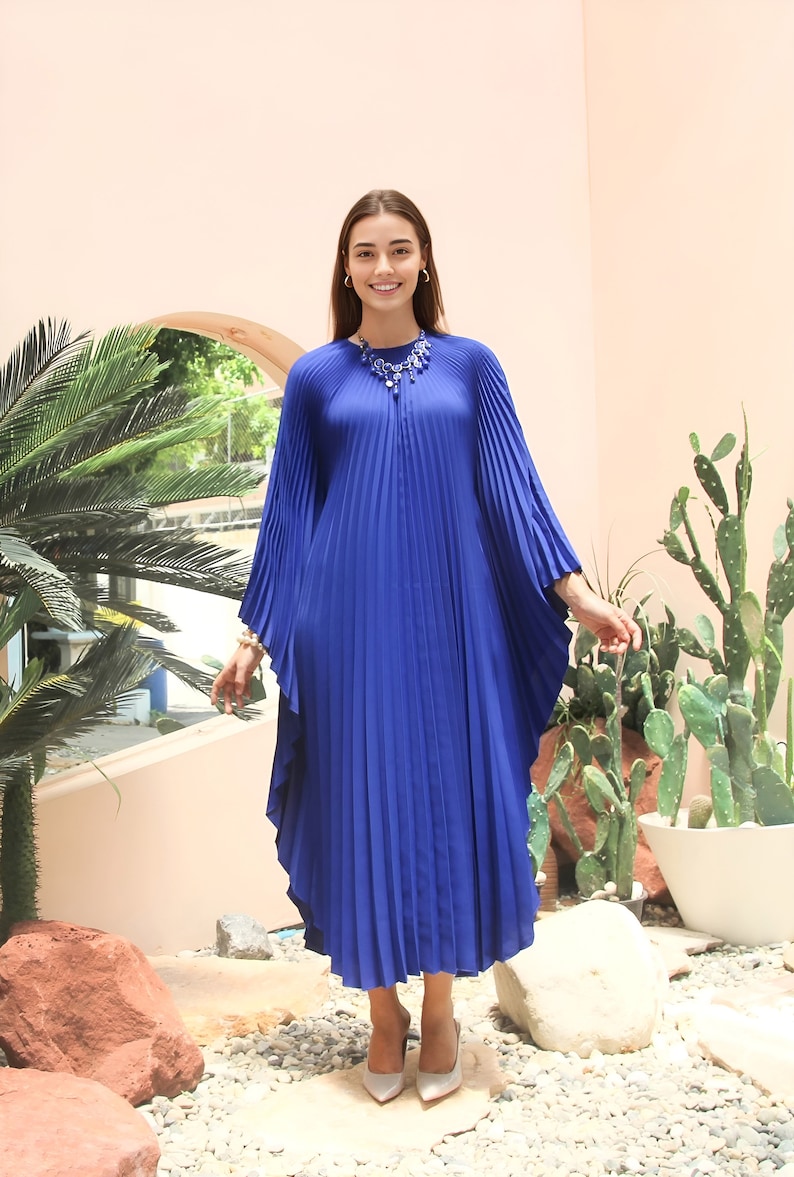 May include: A woman wearing a royal blue pleated maxi dress with long sleeves and a silver necklace. She is standing in front of a pink wall with a cactus and a palm tree in the background.