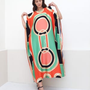 May include: A long, flowing kaftan with a geometric print in shades of orange, green, black, and white. The kaftan has a round neckline and a relaxed fit.