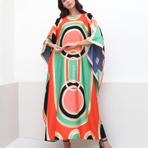May include: A long, flowing kaftan with a geometric print in shades of orange, green, black, and white. The kaftan has a round neckline and wide sleeves.