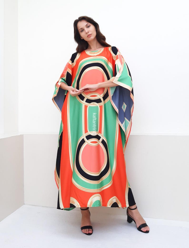 May include: A long, flowing kaftan with a geometric print in shades of orange, green, black, and white. The kaftan has a round neckline and wide sleeves.