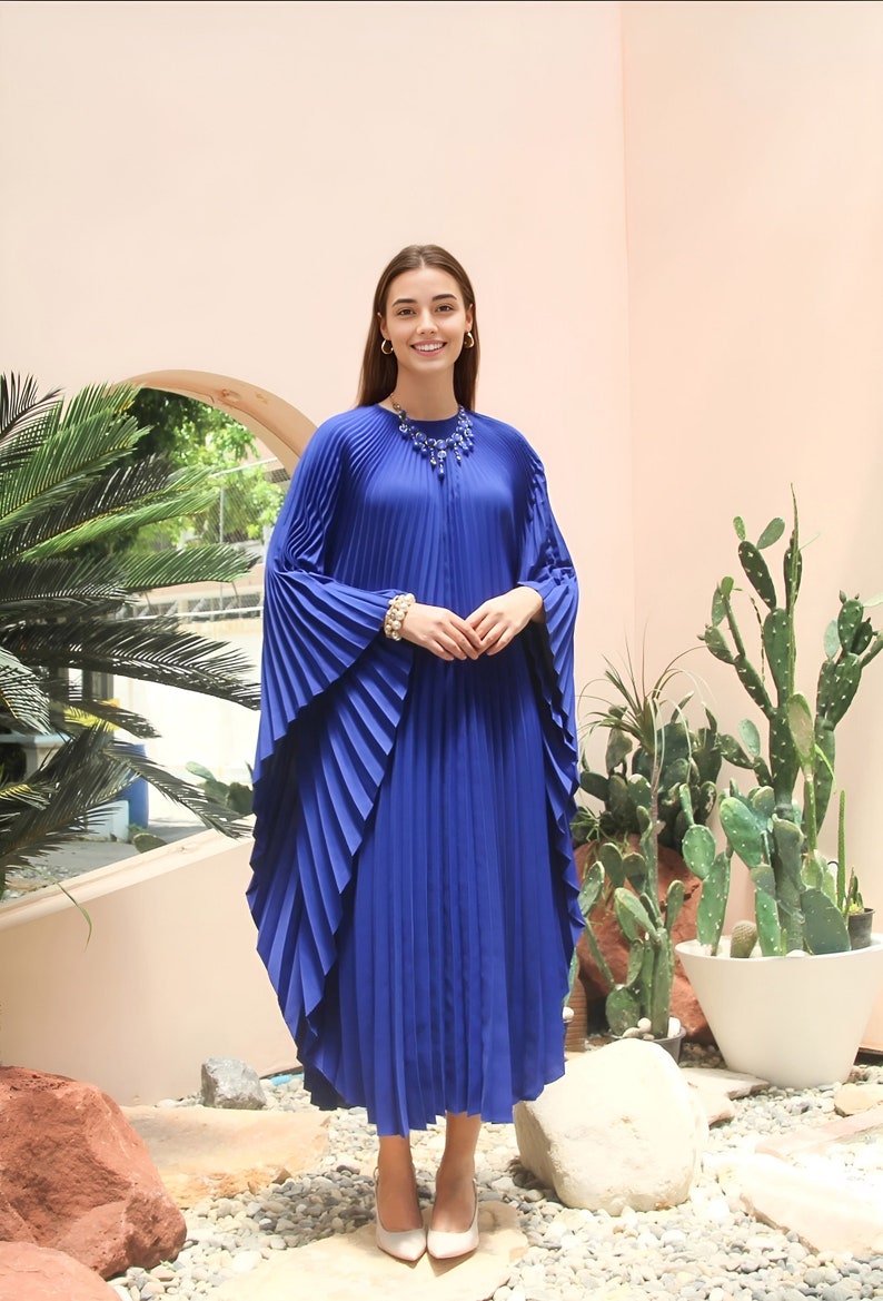 May include: A royal blue pleated maxi dress with long, flowing sleeves. The dress has a round neckline and a beaded necklace.
