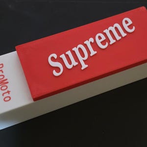 Op de afbeelding: Een rechthoekige doos met een rood deksel en een witte basis. Op het deksel staat het woord "Supreme" in witte, vette letters. De doos is gedeeltelijk open en onthult een wit gedeelte met het woord "ProMoto" in rood.