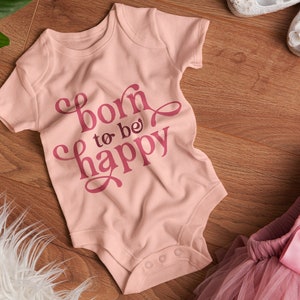 Op de afbeelding: Een lichtroze babyromper met de tekst "born to be happy" in een donkerder roze script. De romper ligt naast een roze tutu, witte schoenen en een wit, pluizig kleed op een houten oppervlak.