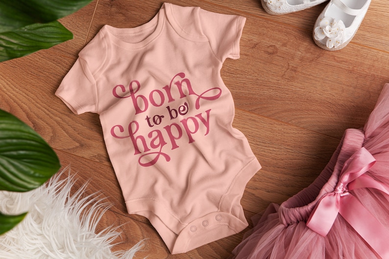 Op de afbeelding: Een lichtroze babyromper met de tekst "born to be happy" in een donkerder roze script. De romper ligt naast een roze tutu, witte schoenen en een wit, pluizig kleed op een houten oppervlak.