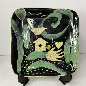 Puede incluir: Un plato cuadrado de cerámica con fondo negro brillante que presenta pinceladas de color verde claro, un pájaro de color crema moteado, una casa, un corazón, un pequeño círculo amarillo y una forma de mano, todo bordeado por un marco color crema.