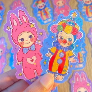 May include: Two die-cut stickers featuring cartoon characters. One is a pink rabbit-costumed figure with a blue bow tie and heart detail. The other is a clown-costumed figure in a yellow and red striped suit with a colorful hat and glitter finish.