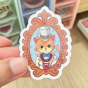 May include: A sticker of a cartoon cat wearing a top hat and bow tie, holding a teacup. The cat is in a gold ornate frame with a blue background.