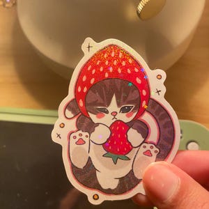 May include: A holographic sticker featuring a brown tabby cat wearing a red strawberry-shaped hood. The cat has dark stripes, pink cheeks, and is holding a small red strawberry with green leaves. The design includes small star and cross accents.