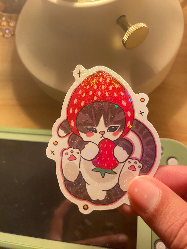 May include: A holographic sticker featuring a brown tabby cat wearing a red strawberry-shaped hood. The cat has dark stripes, pink cheeks, and is holding a small red strawberry with green leaves. The design includes small star and cross accents.