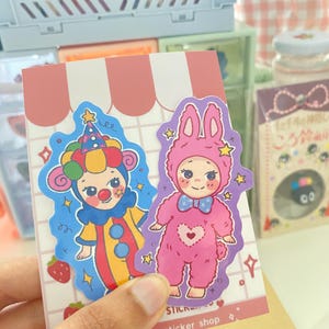May include: Two die-cut stickers on a white card with pink stripes. One sticker features a clown in a colorful yellow, red, and blue outfit with a matching hat. The other sticker shows a character in a pink fuzzy bunny suit with a blue bow tie.