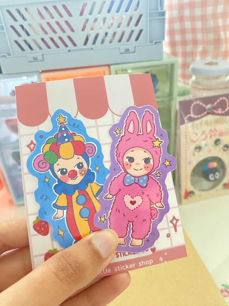 May include: Two die-cut stickers on a white card with pink stripes. One sticker features a clown in a colorful yellow, red, and blue outfit with a matching hat. The other sticker shows a character in a pink fuzzy bunny suit with a blue bow tie.