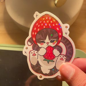 May include: A holographic sticker featuring a brown and white tabby cat wearing a red strawberry-shaped hood. The cat is depicted holding a small red strawberry with green leaves. The design includes small star and cross accents on a glittery finish.