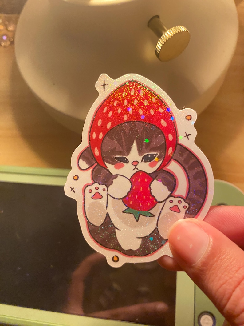 May include: A holographic sticker featuring a brown and white tabby cat wearing a red strawberry-shaped hood. The cat is depicted holding a small red strawberry with green leaves. The design includes small star and cross accents on a glittery finish.