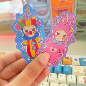 May include: Two die-cut stickers featuring cartoon characters. One sticker shows a clown with a red nose, yellow and red outfit, and a blue hat. The other sticker depicts a character in a pink fuzzy rabbit costume with a blue bow tie and heart.
