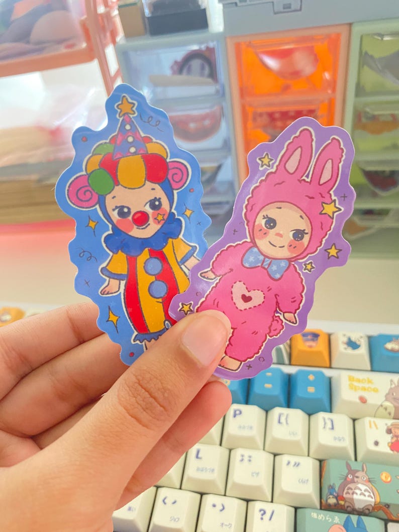 May include: Two die-cut stickers featuring cartoon characters. One sticker shows a clown with a red nose, yellow and red outfit, and a blue hat. The other sticker depicts a character in a pink fuzzy rabbit costume with a blue bow tie and heart.