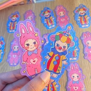 May include: Die-cut stickers featuring two distinct character designs: a pink bunny-costumed figure with a heart motif and a colorful clown figure with a polka-dot hat. Both stickers have a holographic glitter finish and star accents.