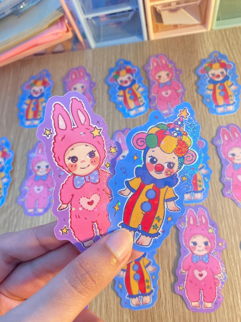 May include: Die-cut stickers featuring two distinct character designs: a pink bunny-costumed figure with a heart motif and a colorful clown figure with a polka-dot hat. Both stickers have a holographic glitter finish and star accents.