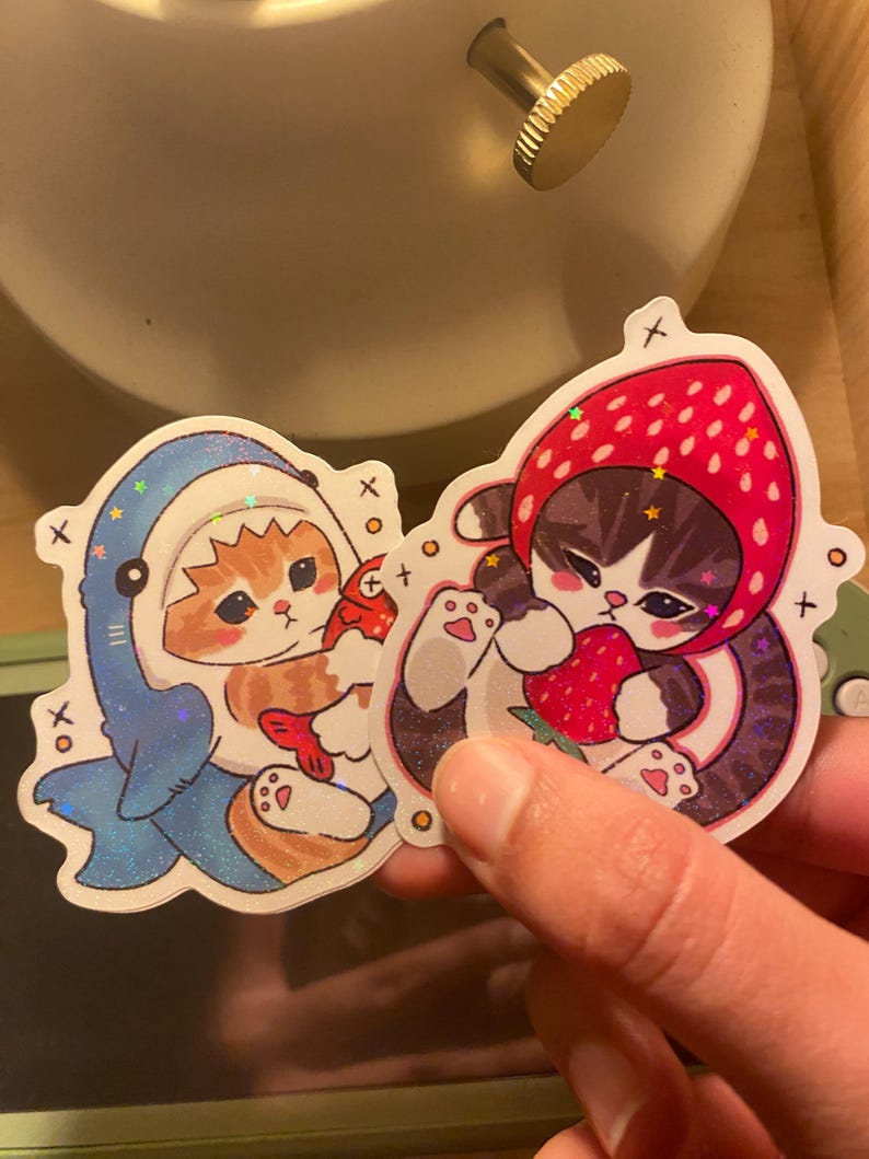 May include: Two die-cut stickers featuring illustrated cats. One sticker shows an orange tabby cat wearing a blue shark costume holding a red fish. The other sticker shows a grey and white cat wearing a red strawberry hood holding a strawberry.