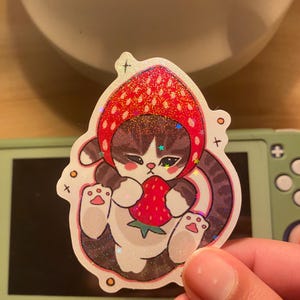 May include: A die-cut sticker featuring a brown tabby cat wearing a red strawberry-shaped hood. The cat is holding a small red strawberry with green leaves. The sticker has a holographic finish with small star-shaped sparkles throughout the design.