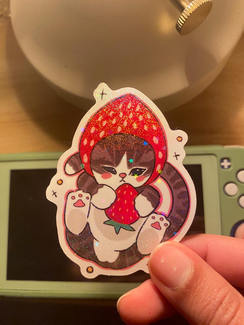 May include: A die-cut sticker featuring a brown tabby cat wearing a red strawberry-shaped hood. The cat is holding a small red strawberry with green leaves. The sticker has a holographic finish with small star-shaped sparkles throughout the design.