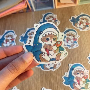 May include: A die-cut sticker featuring a brown tabby cat wearing a blue shark costume. The cat is holding a small, colourful fish. The sticker has a holographic finish with small star accents and black 'x' and 'o' symbols printed on the sides.